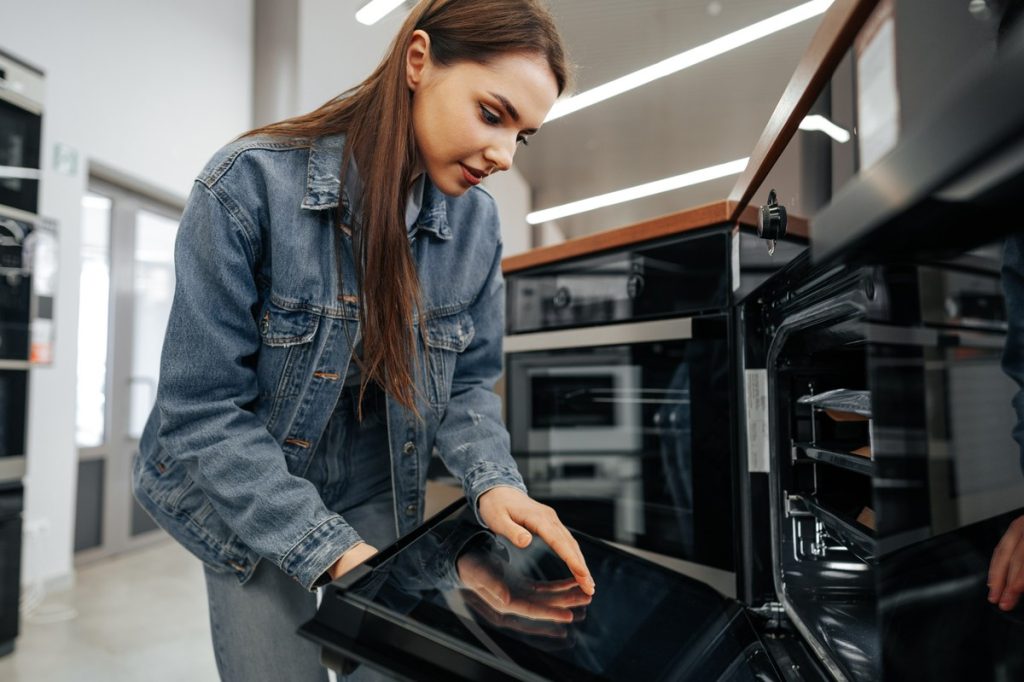 Une jeune femme examine attentivement un four moderne dans un magasin d’électroménager, en ouvrant la porte pour observer l’intérieur.