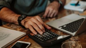 Un homme utilise une calculatrice financière sur une table en bois à côté d’un ordinateur portable pour planifier ses économies et ses investissements.